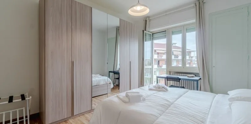 Un dormitorio luminoso con una cama grande, un armario y ventanas frente a edificios cercanos.