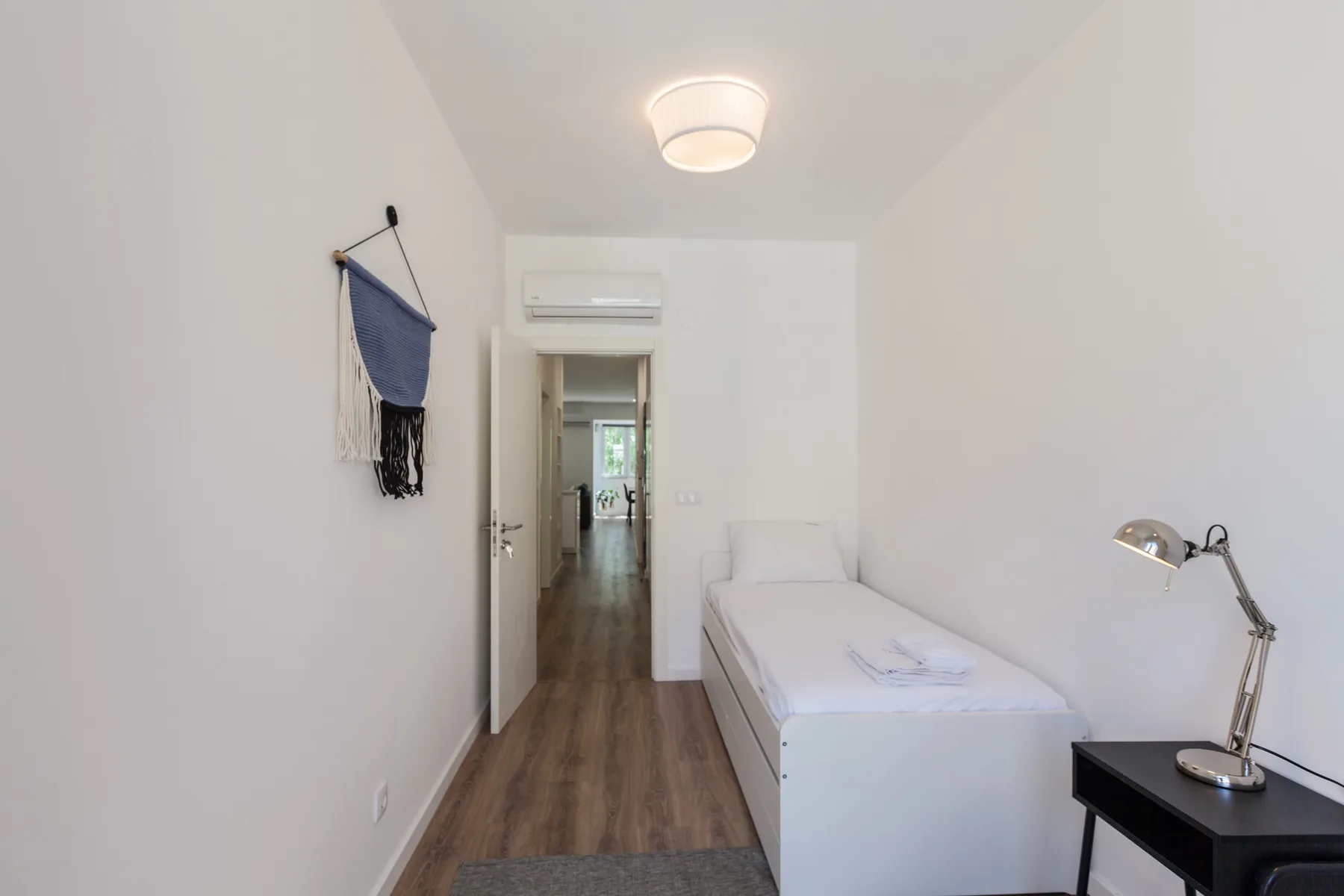 Un dormitorio estrecho con una cama individual, una mesa auxiliar y una lámpara de escritorio.
