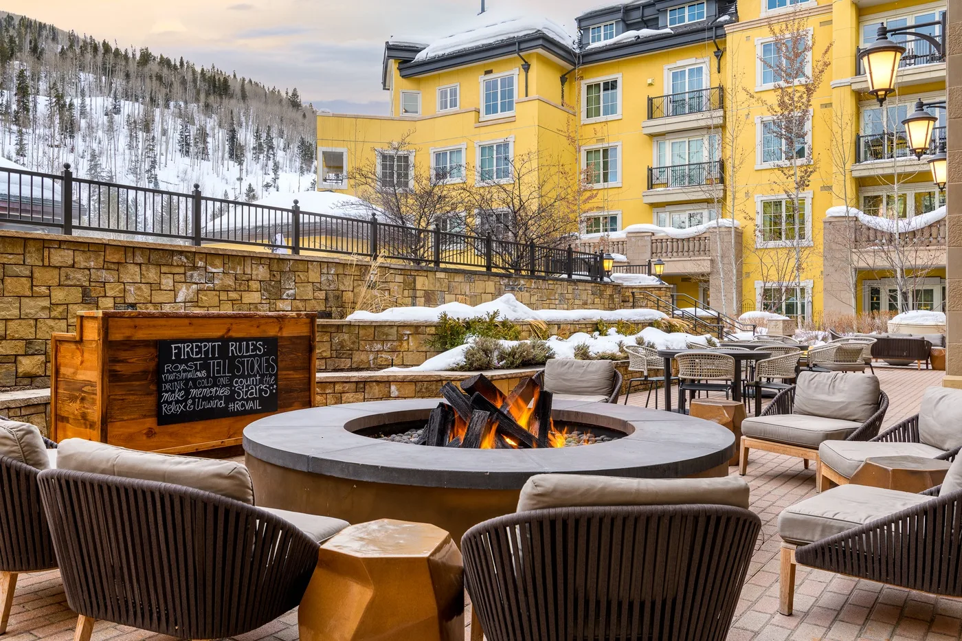 An outdoor patio with a circular fire pit and seating beside a yellow building.