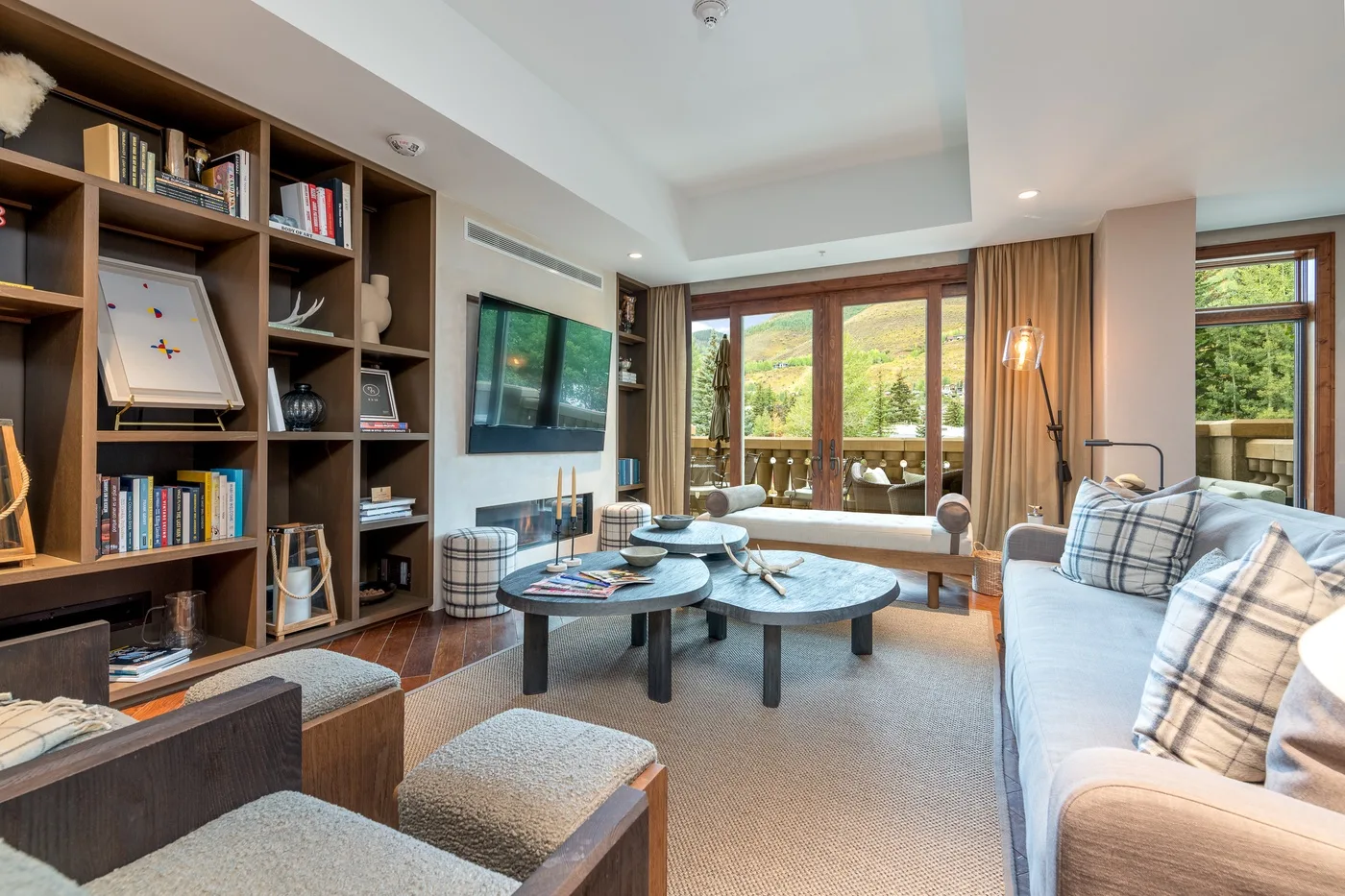 A bright living room with seating, a wall-mounted television, and large windows overlooking greenery.