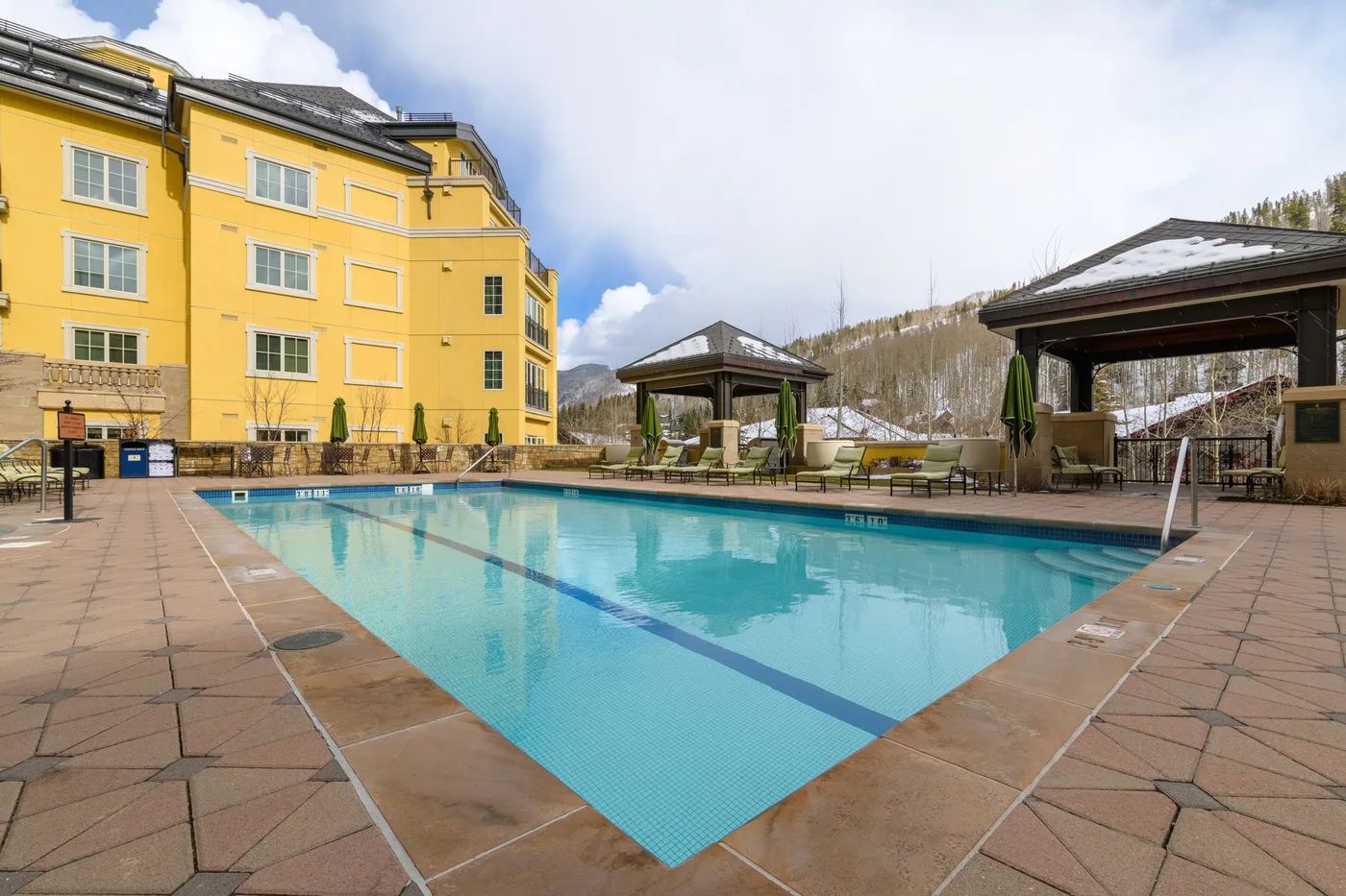 An outdoor swimming pool beside a yellow multi-story building and covered seating areas.