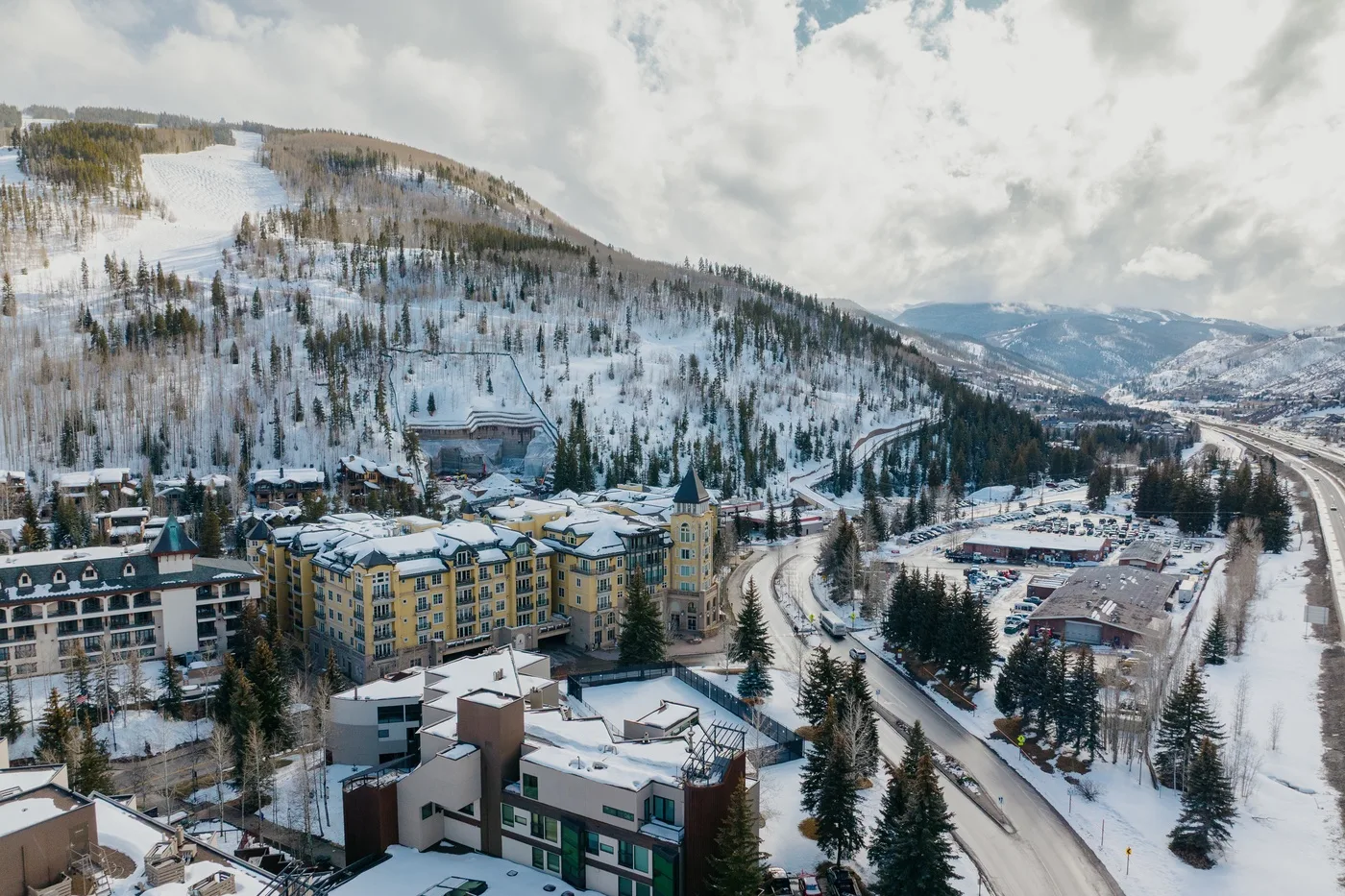 Snow-covered buildings sit below a wooded mountain slope in a winter resort scene.