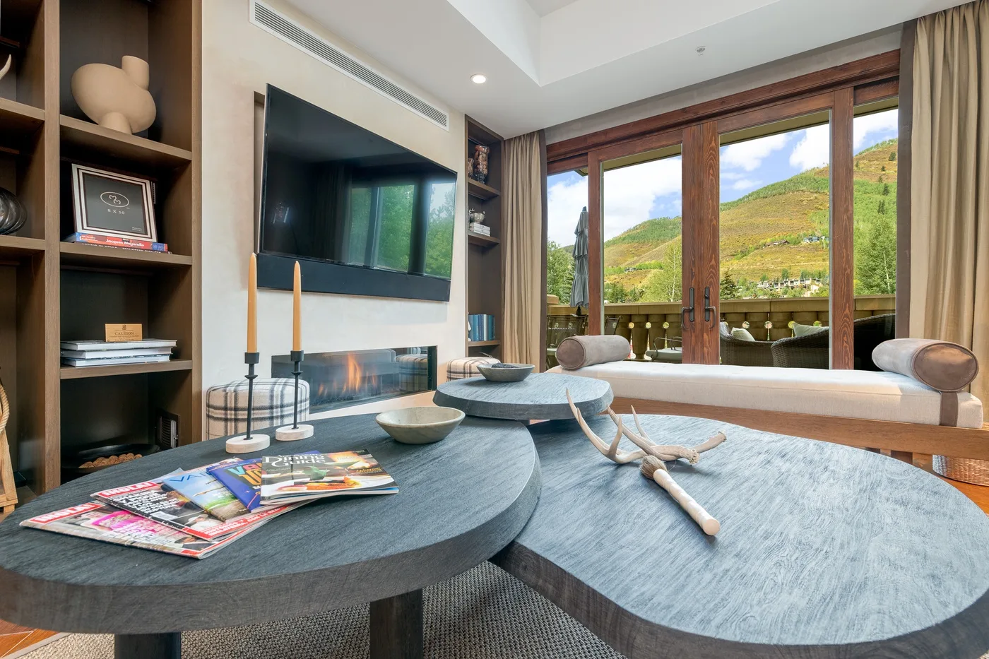 A living room with a television, low tables, and a wide window overlooking green hills.