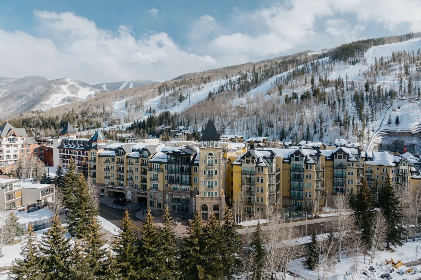 Snow-covered resort buildings sit below a wooded mountain slope under a partly cloudy sky.