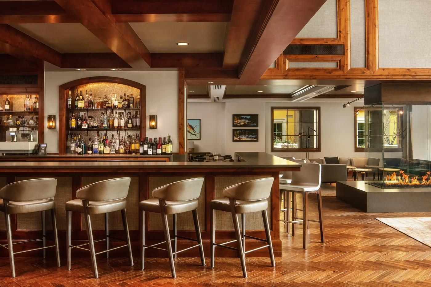 A warmly lit bar with a long counter, stools, and bottles on display.
