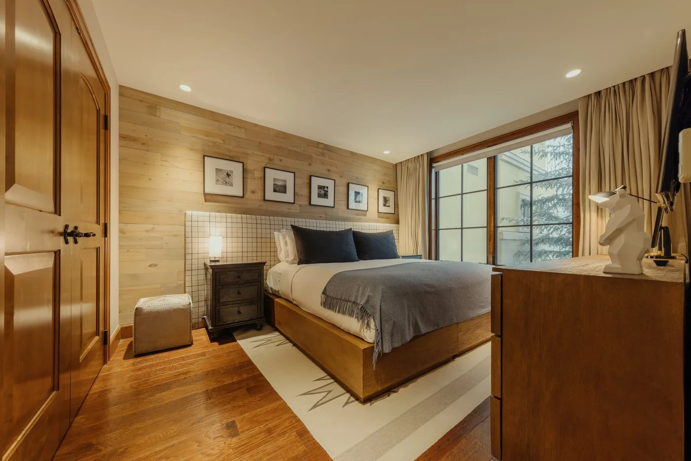 A bedroom with a wooden bed, bedside table, and large curtained window.