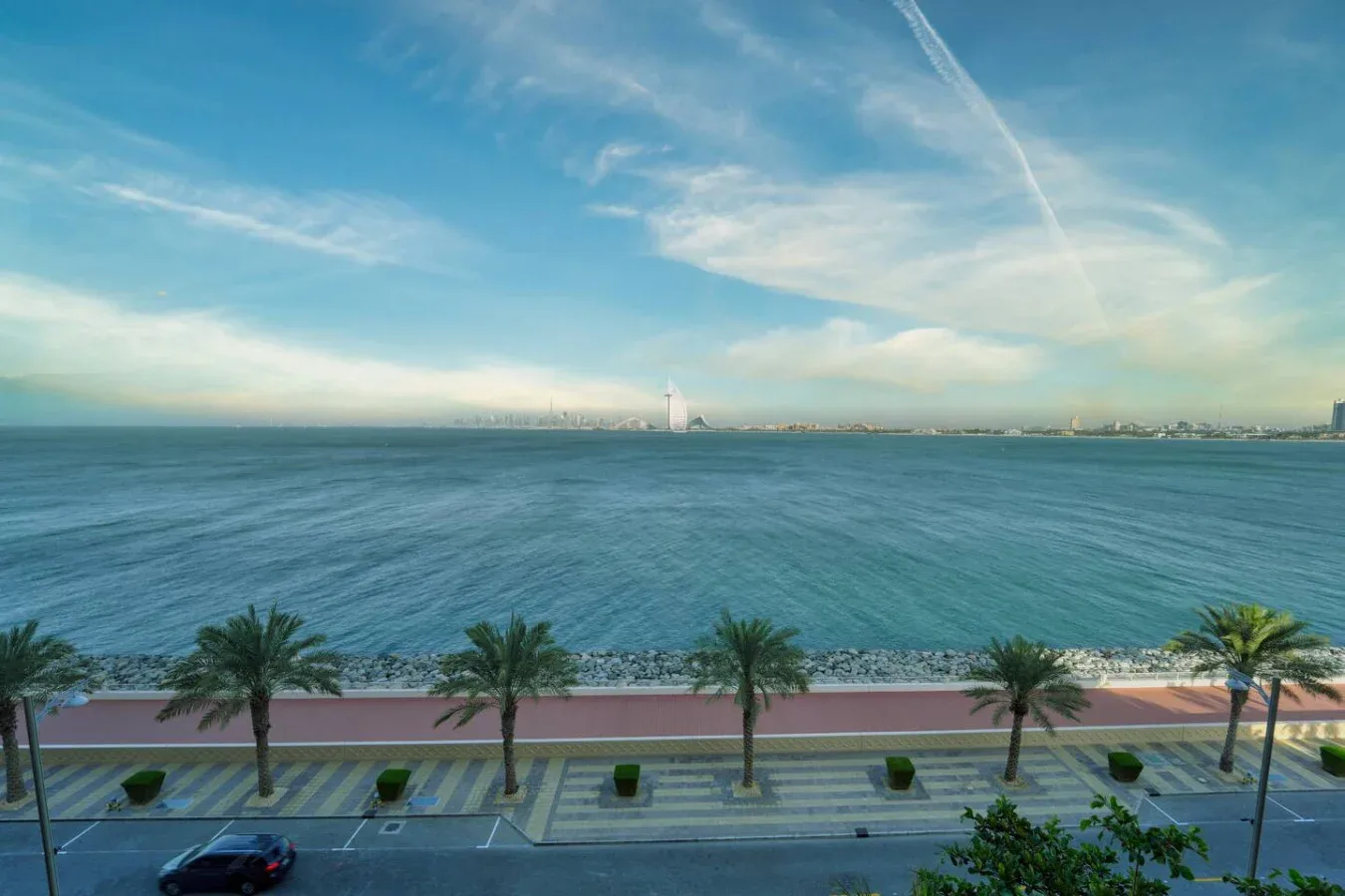 Vista frente al mar hacia un perfil urbano lejano, con palmeras a lo largo del paseo.