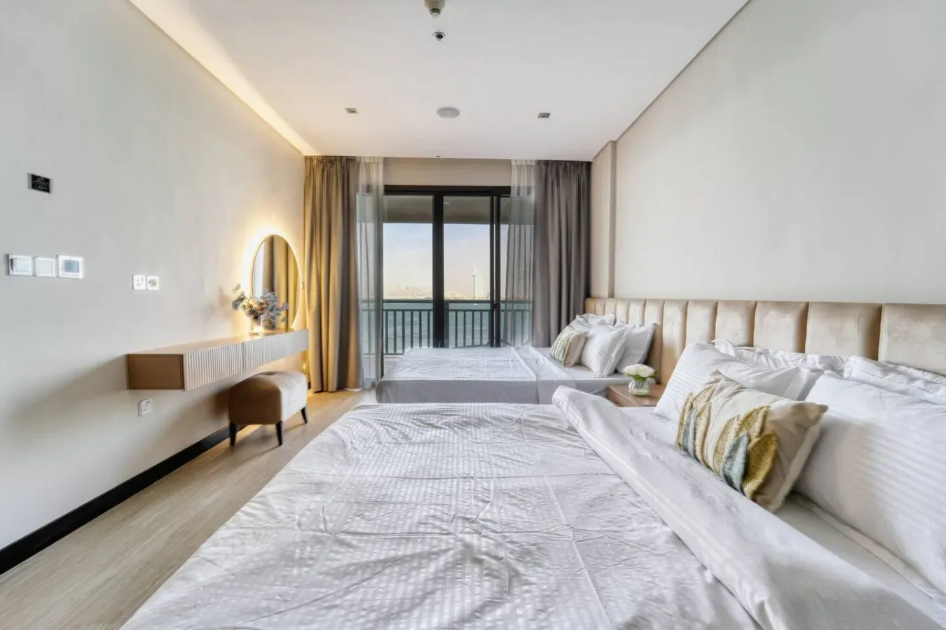 Un dormitorio luminoso con ropa de cama blanca, un tocador y puertas de vidrio hacia un balcón.