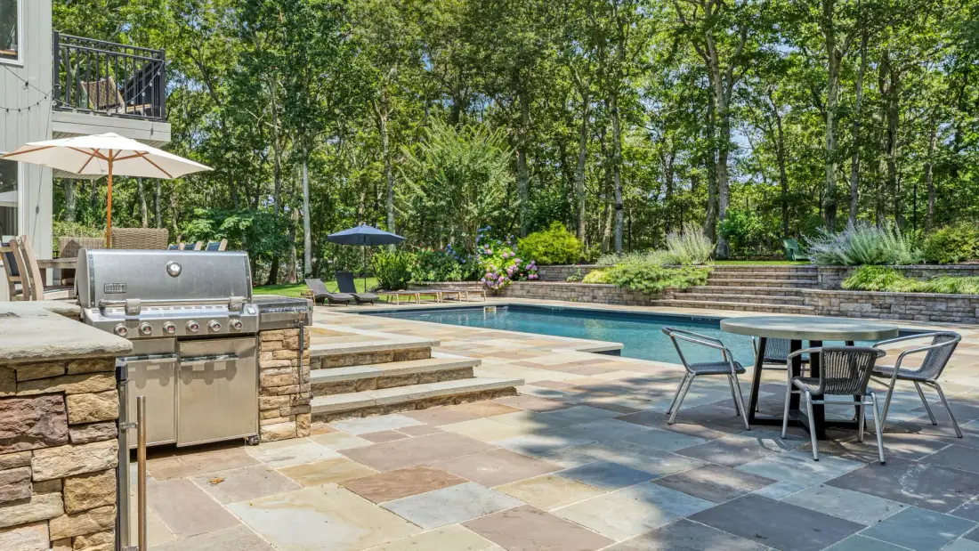 Una zona de piscina al aire libre con patio pavimentado, mesa de comedor y parrilla.