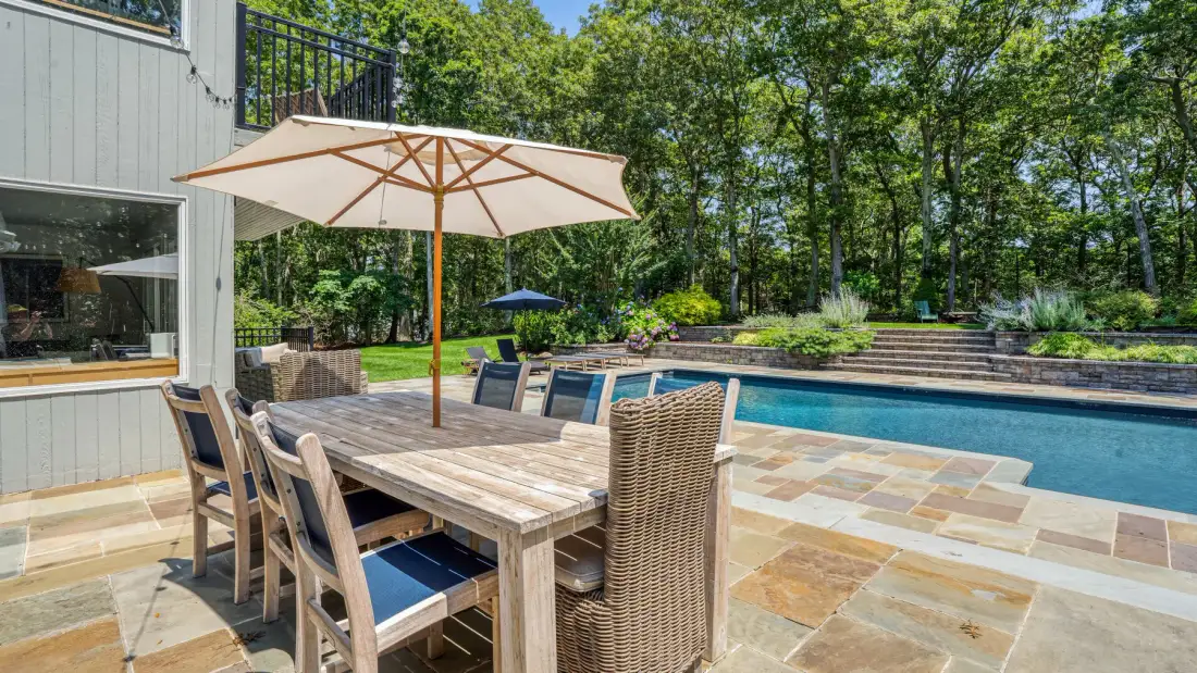 Una zona de comedor al aire libre junto a una piscina, con una sombrilla grande sobre la mesa.