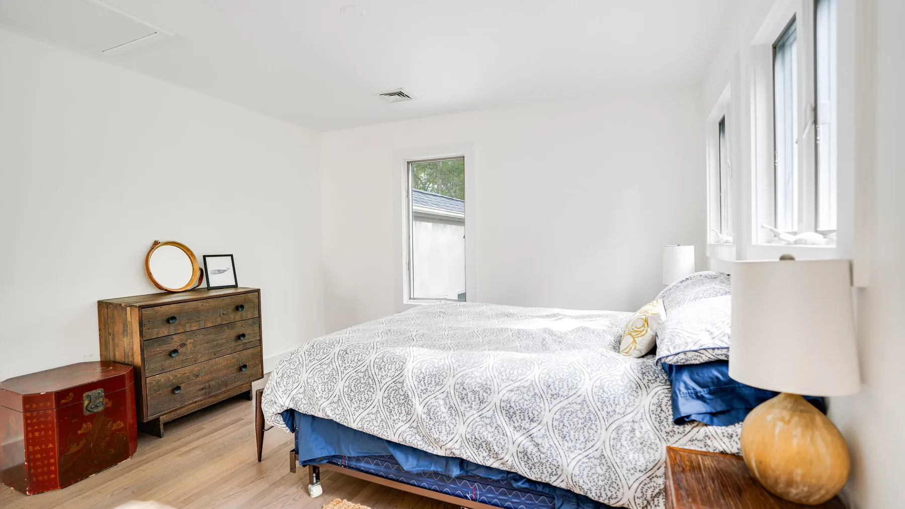 Un dormitorio luminoso con una cama, una cómoda de madera y ventanas grandes.