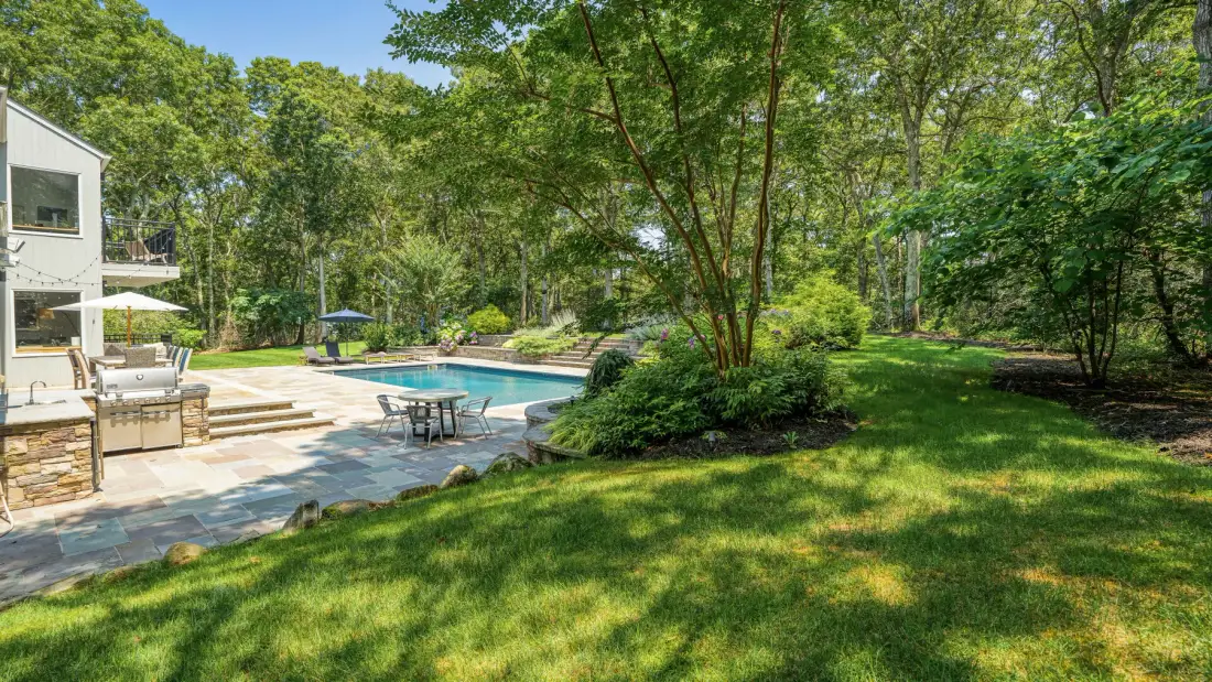 Un patio ajardinado con piscina, asientos y árboles frondosos.