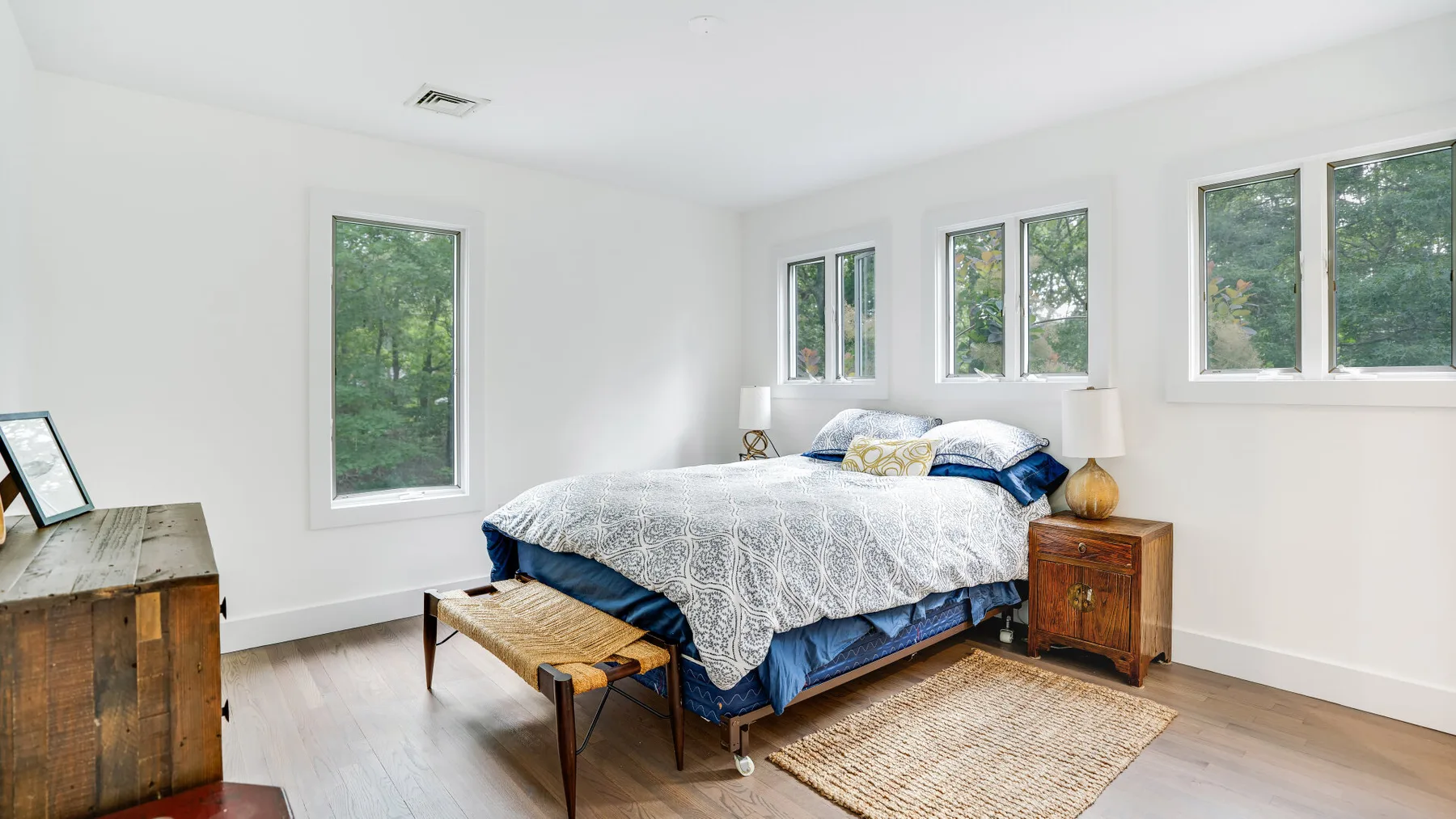 Un dormitorio luminoso con una cama, mesitas de noche y ventanas con vistas a los árboles.
