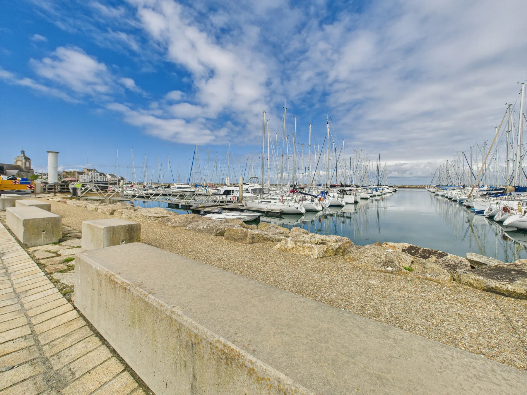 A marina with sailboats sits beside a stone waterfront promenade under a partly cloudy sky.