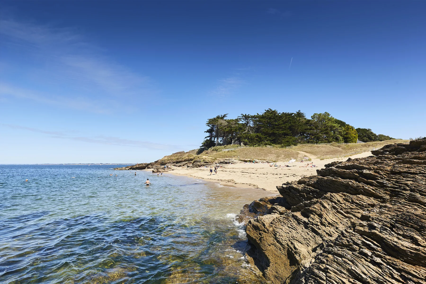 A rocky shoreline borders a sandy beach and calm blue water.