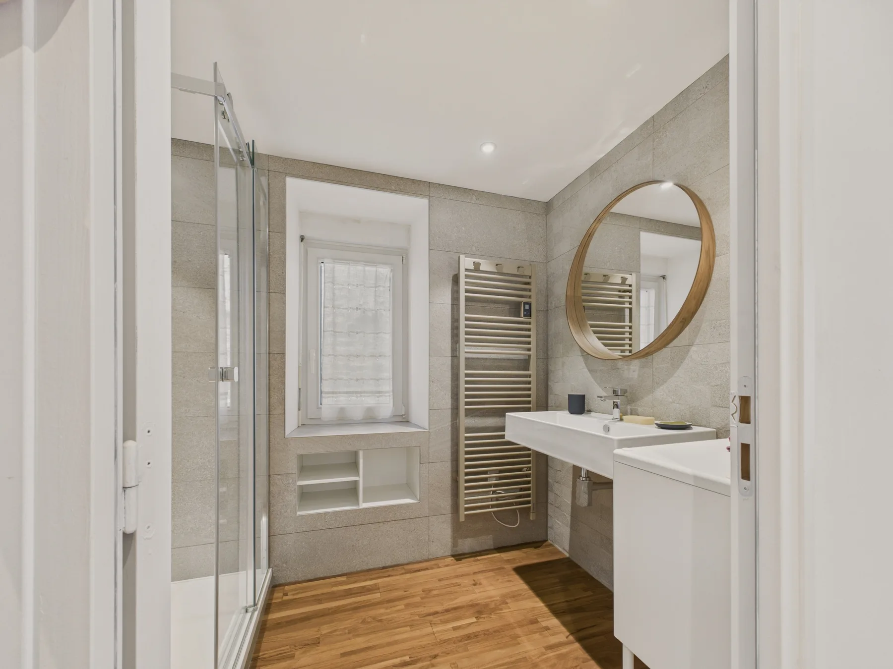 A bathroom with a glass shower, vanity, and round mirror.