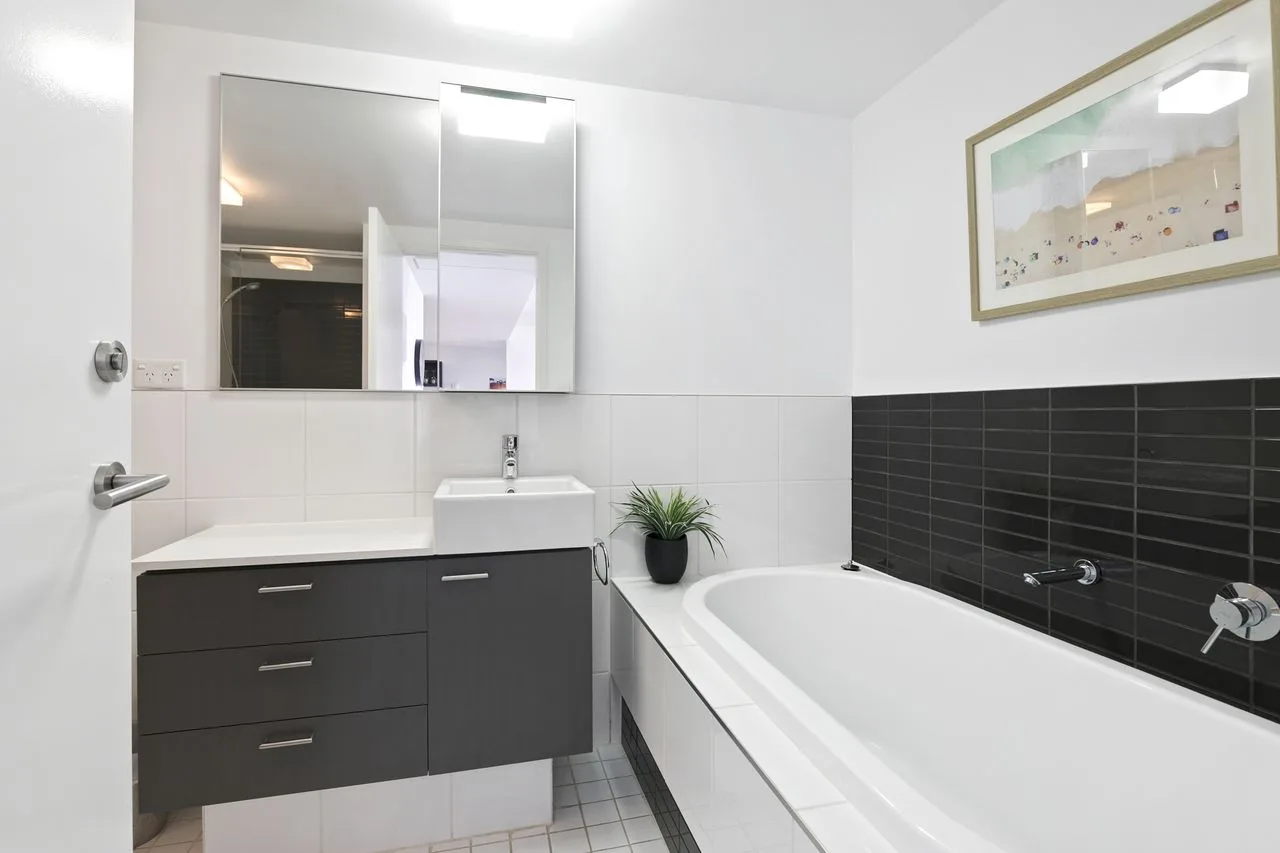 A bright bathroom with a bathtub, dark tiled wall, and vanity.