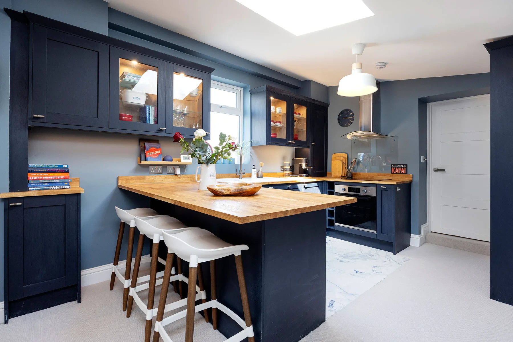 A dark blue kitchen with a wooden island and white bar stools.