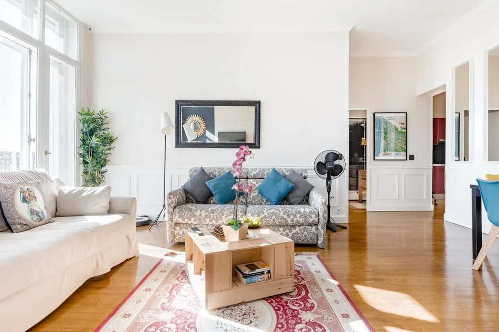 A bright living room with patterned seating, a wooden coffee table, and large windows.