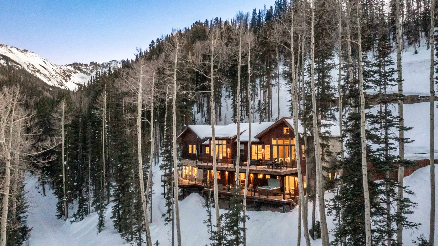 A warmly lit lodge sits among snow-covered trees on a mountain slope.