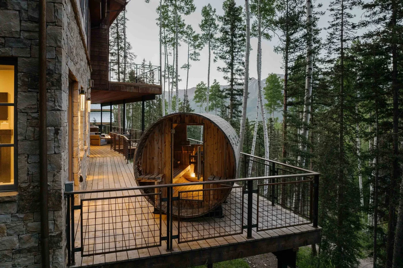 A wooden deck beside a stone building overlooks a forest, with a rounded wooden structure.