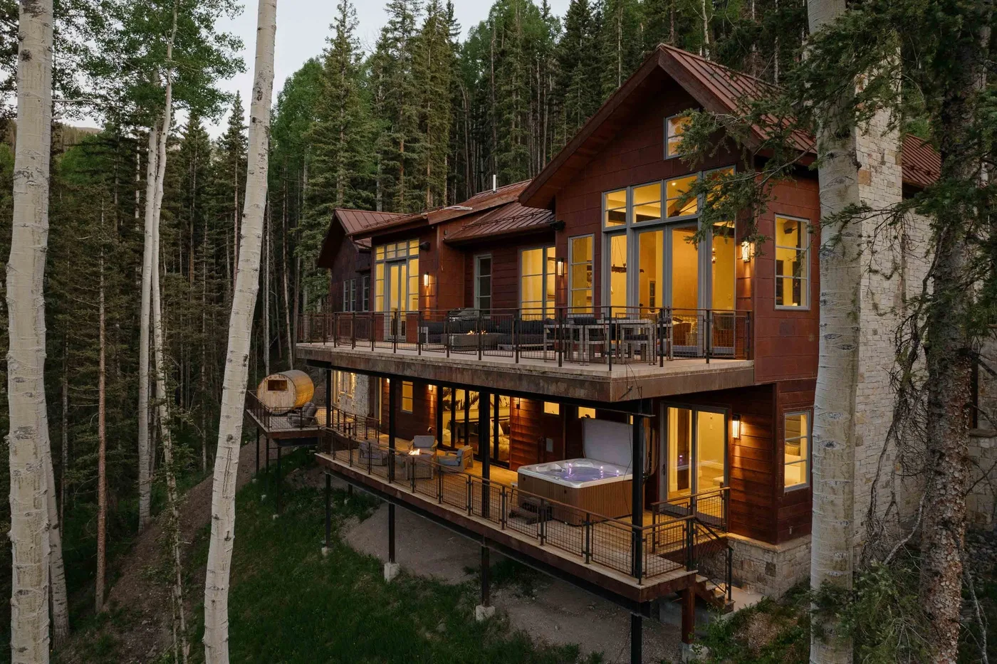 A modern cabin nestled among trees, with a large deck and glowing windows.