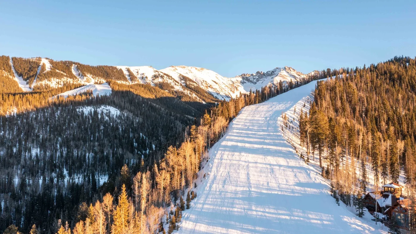 A snow-covered ski slope runs between forested mountain ridges beneath a clear sky.