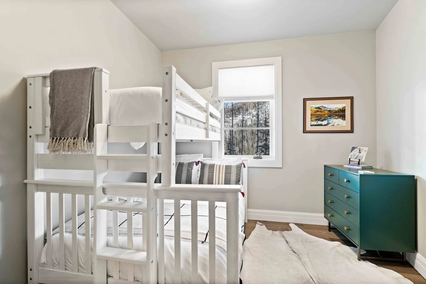 A bright bedroom with white bunk beds, a window, and a teal dresser.