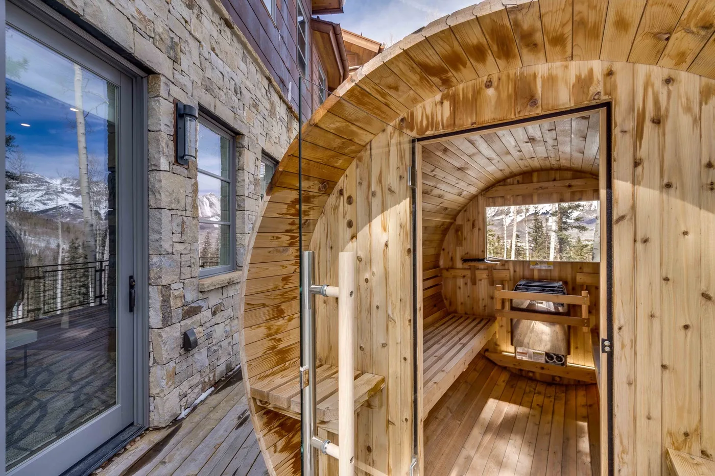 A wooden outdoor sauna with a small seating area beside a stone-clad building.