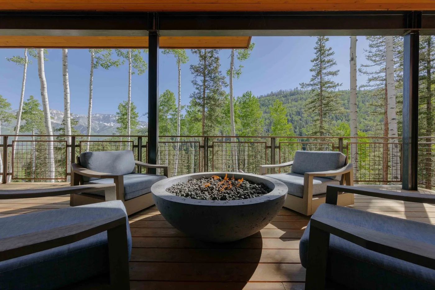 A furnished outdoor deck with a round fire pit overlooks a wooded hillside.