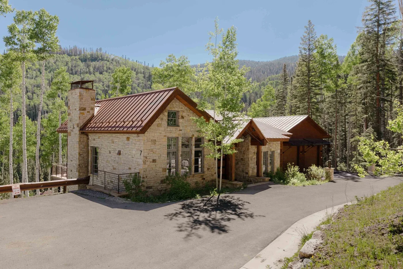 A stone house with a metal roof sits beside a paved driveway in a wooded mountain setting.