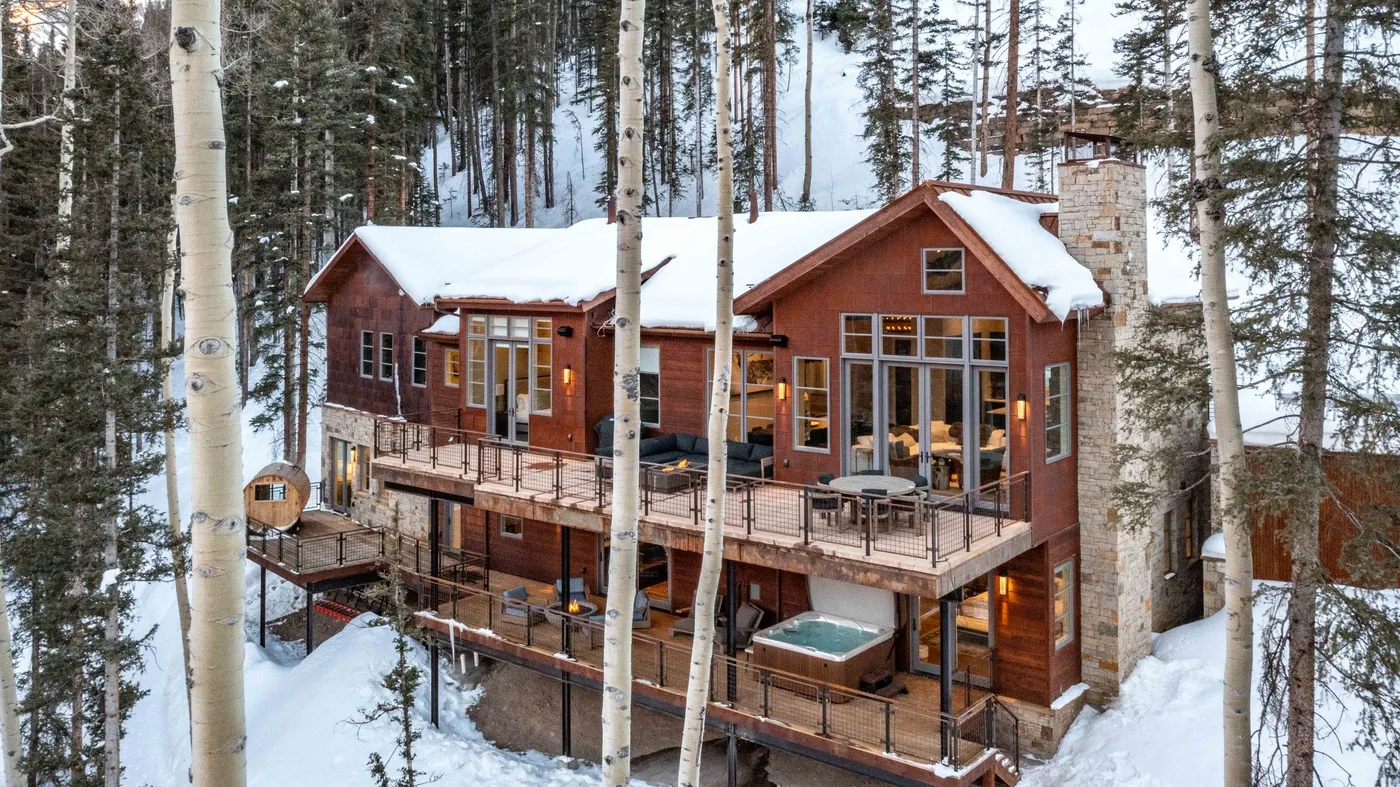 A large wooden cabin with wraparound decks stands among snow-covered trees.
