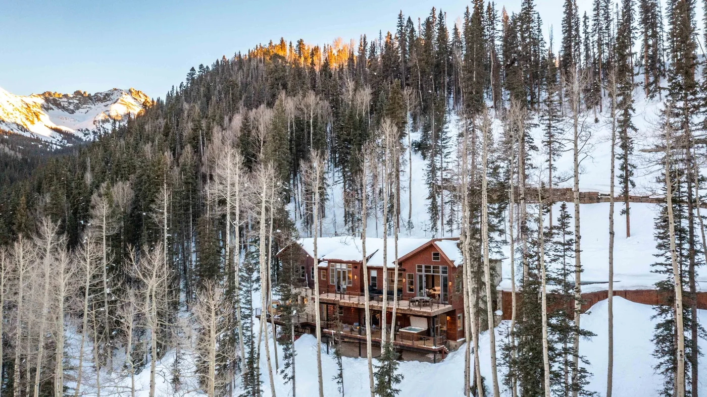 A wooded mountain cabin stands among snow-covered trees beneath a forested ridge.