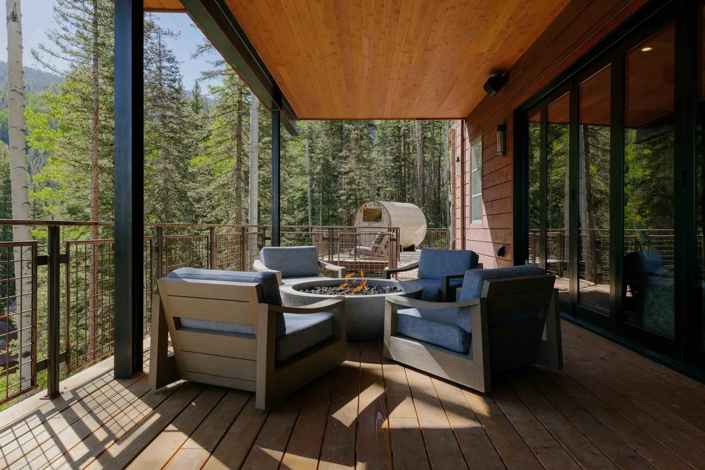 A covered outdoor seating area with a fire pit overlooks a wooded landscape.