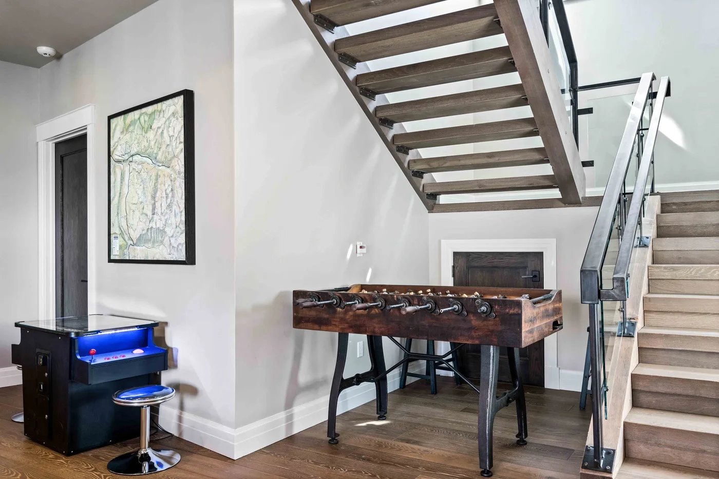 A foosball table sits beneath an open staircase in a bright room.