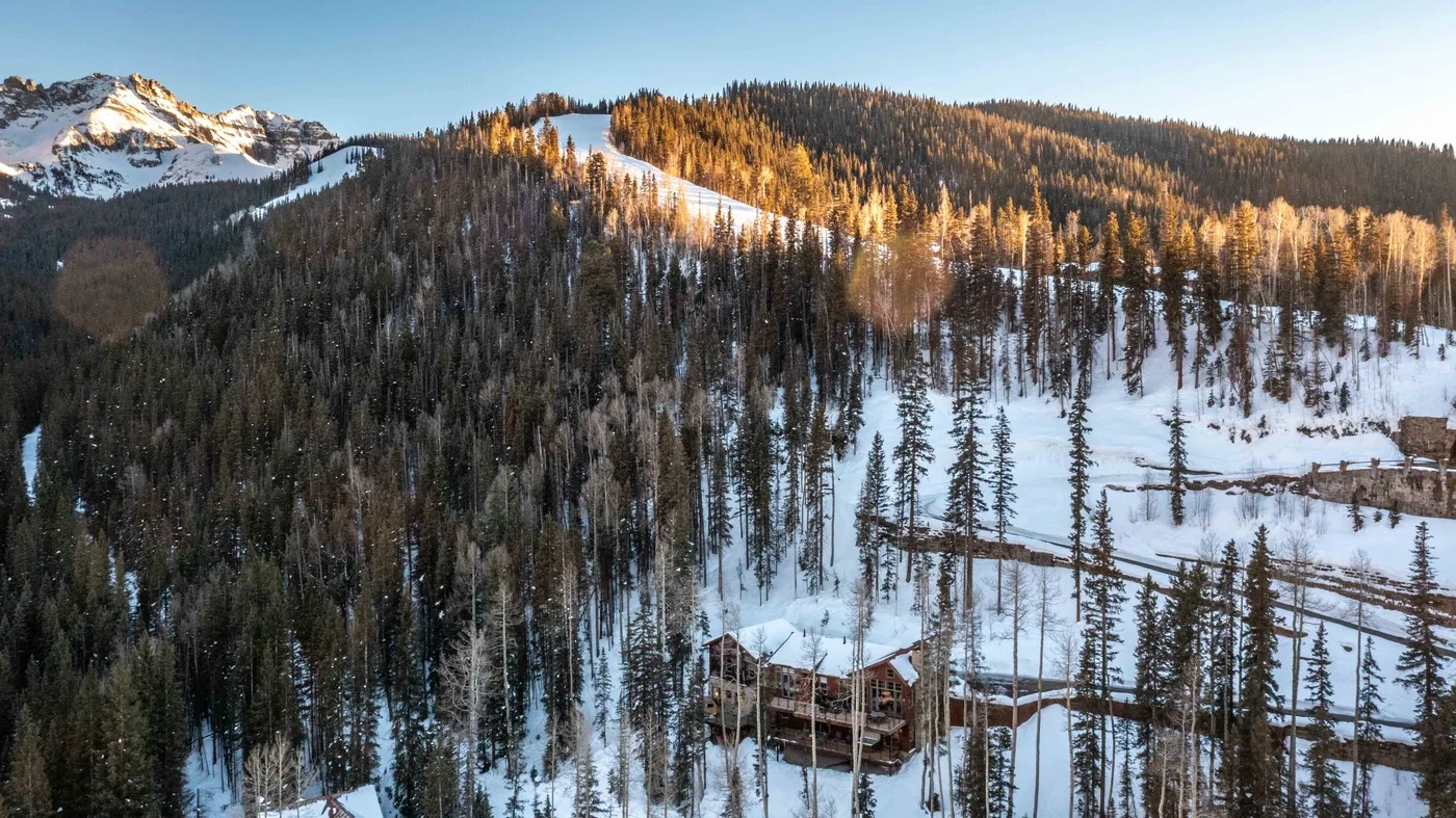 A snowy mountain forest surrounds a small cabin on the hillside.