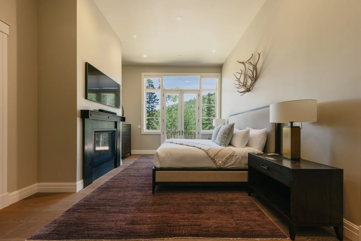 A bedroom with a bed, bedside table, and large windows overlooking trees.