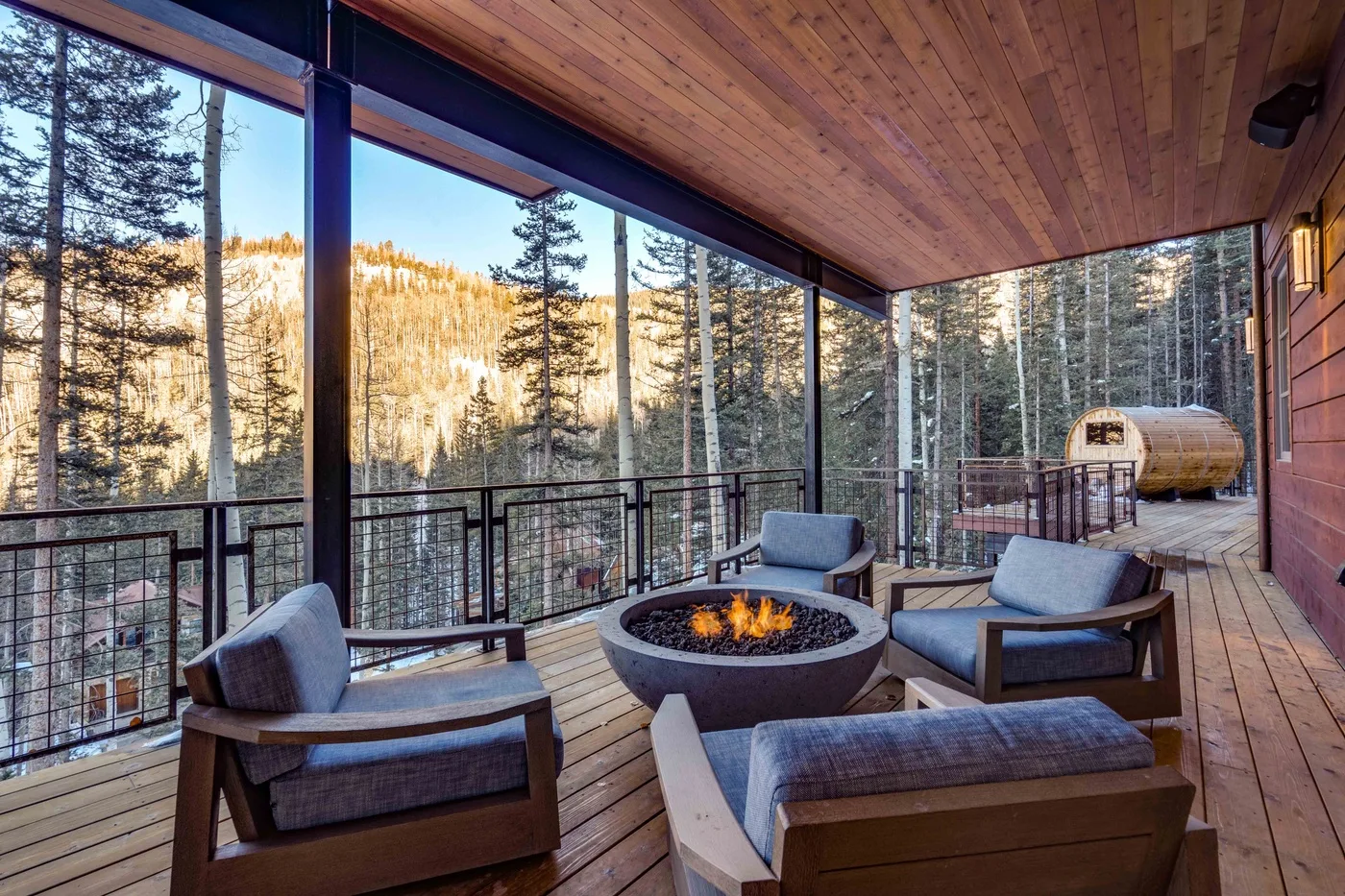A covered wooden deck with outdoor seating and a fire pit overlooks a forested hillside.