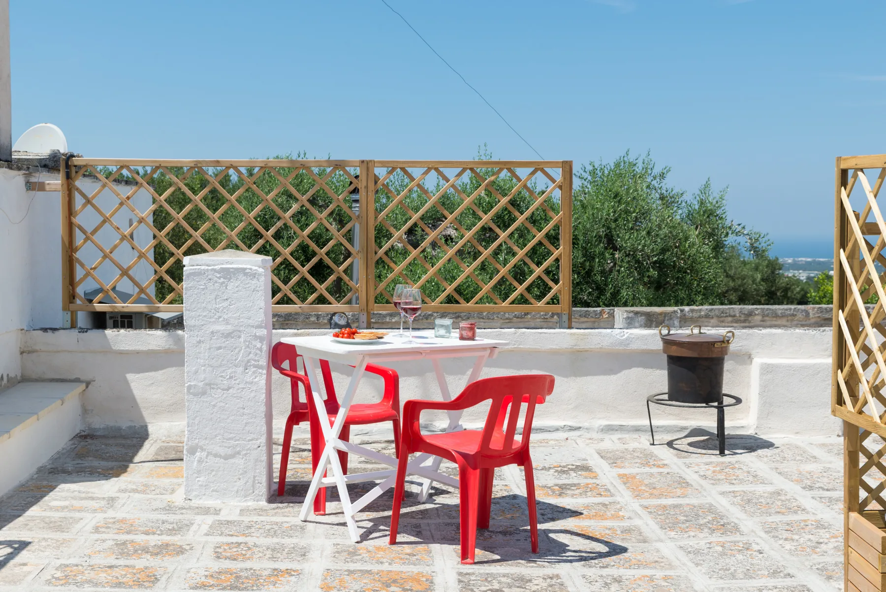 Una terraza en la azotea tiene una mesa blanca y asientos rojos junto a una celosía de madera.
