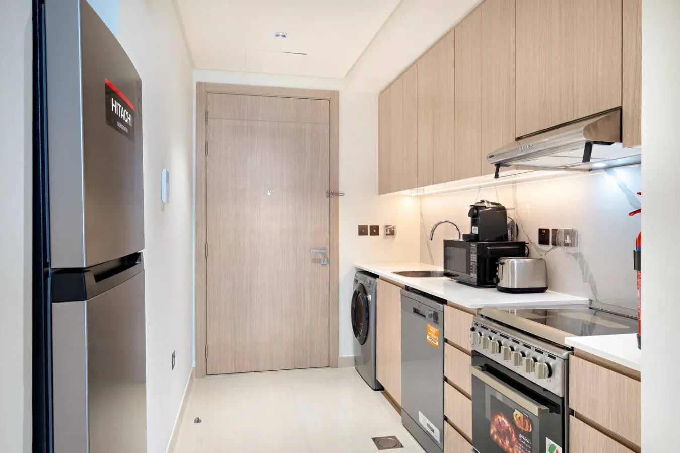 A narrow kitchen with light wood cabinets, appliances, and a closed door.