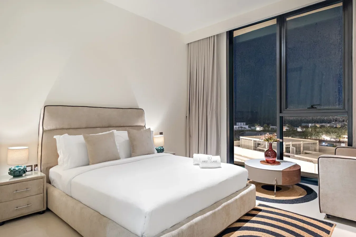 A bedroom with a neatly made bed beside large windows and a seating area.