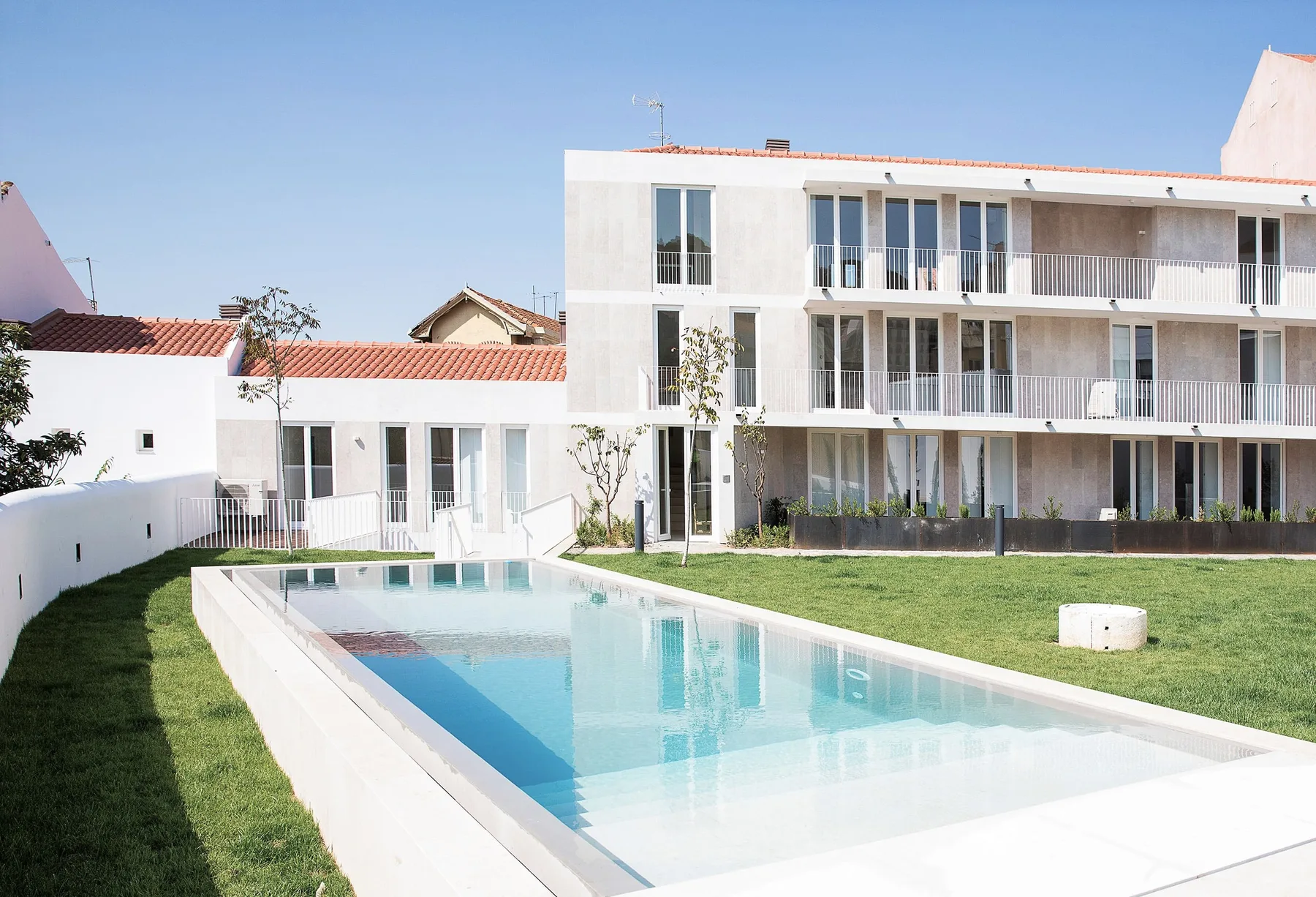 A swimming pool sits beside a grassy courtyard and modern white buildings.
