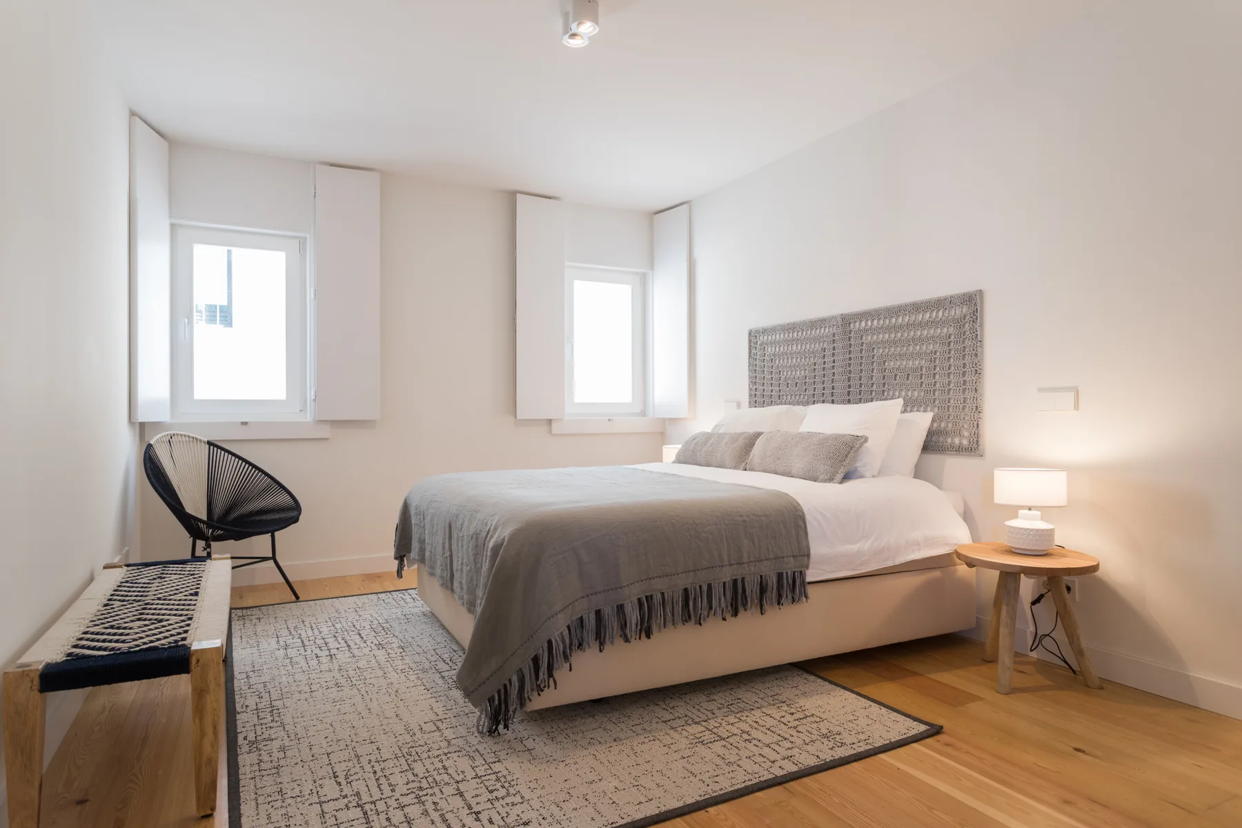 A bright bedroom with a large bed, two windows, and a bedside table.