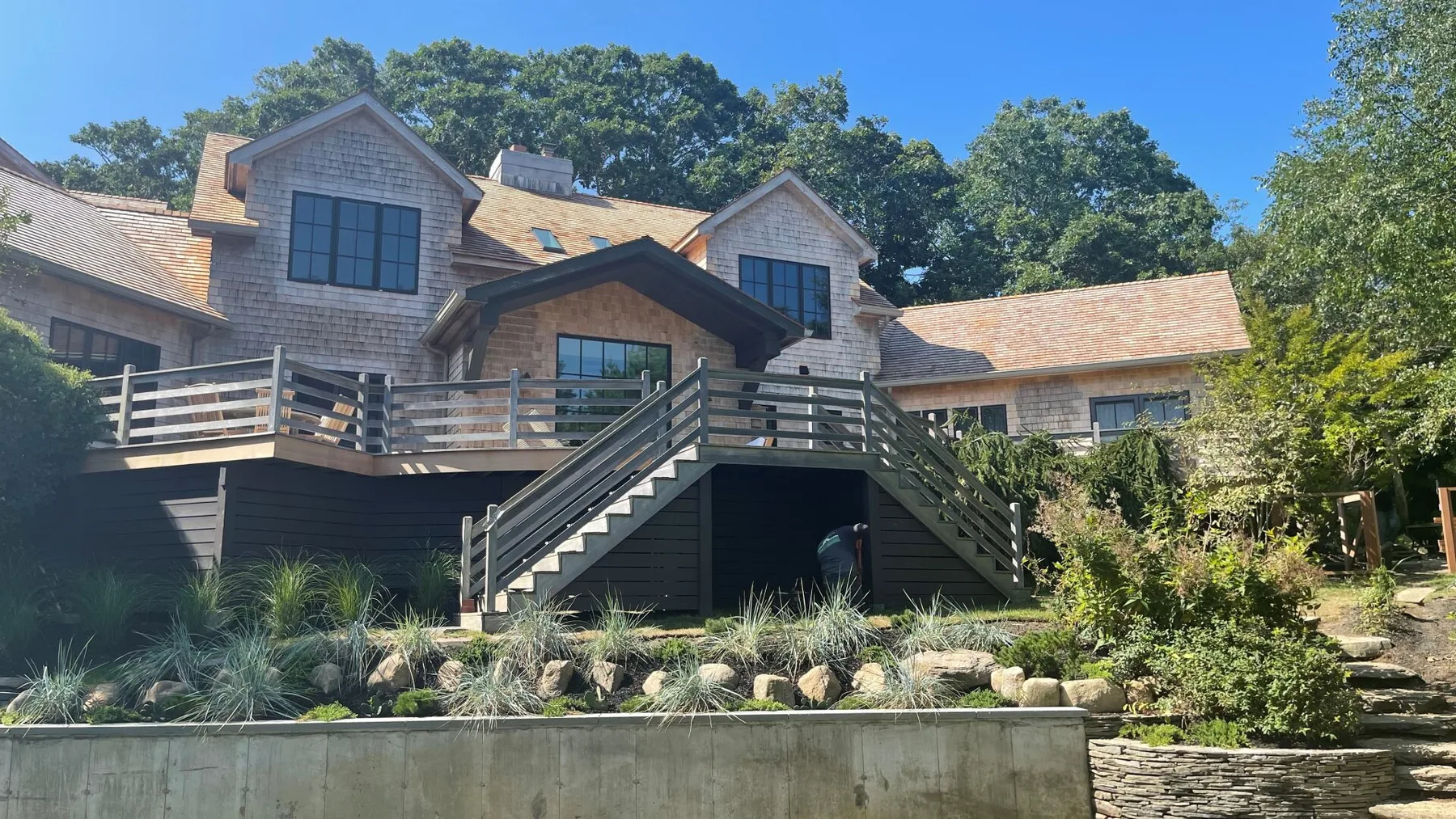 A large house with a raised wooden deck and central staircase sits among trees.