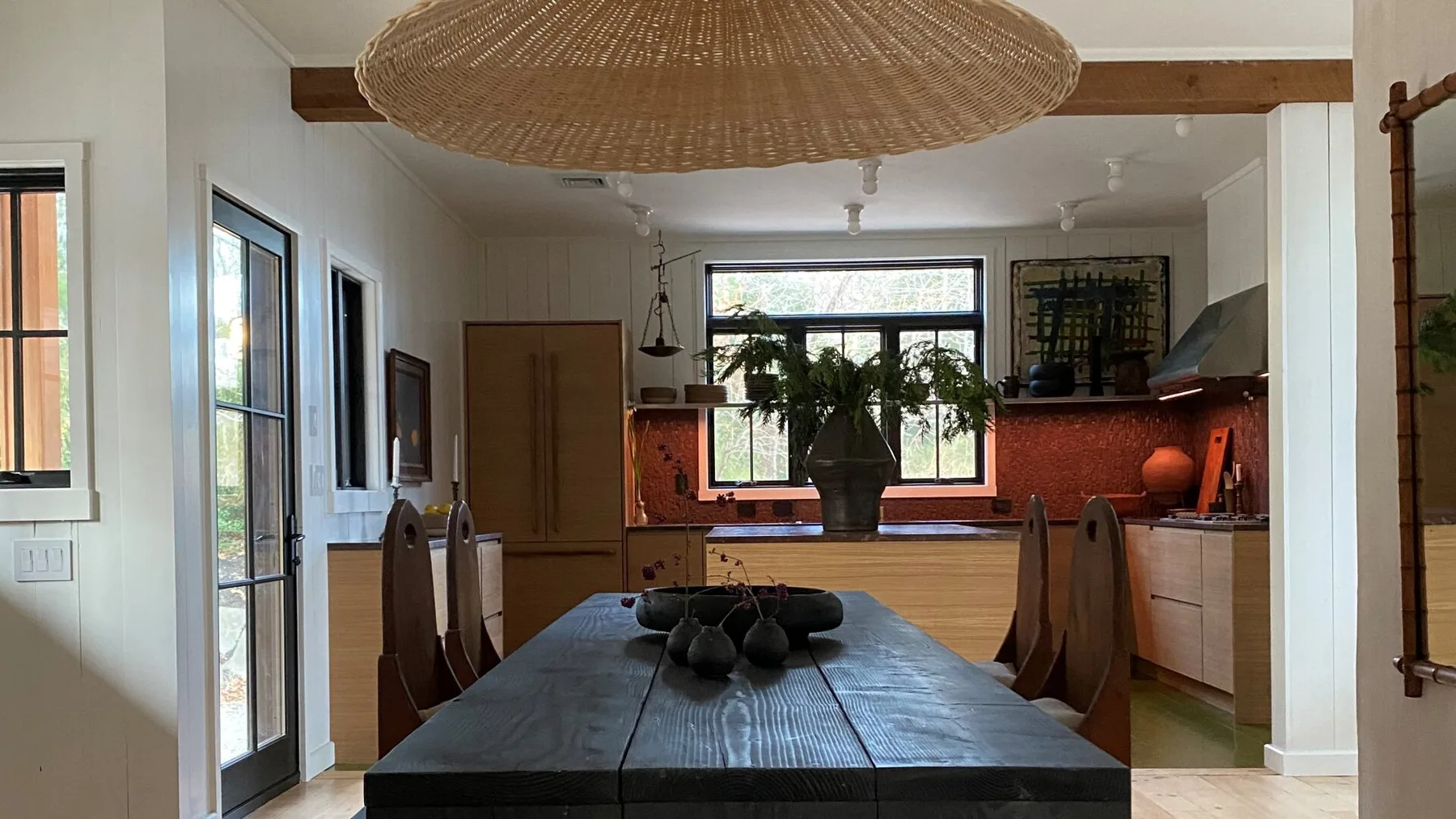 A dining area opens onto a kitchen, with a long table beneath a woven pendant light.