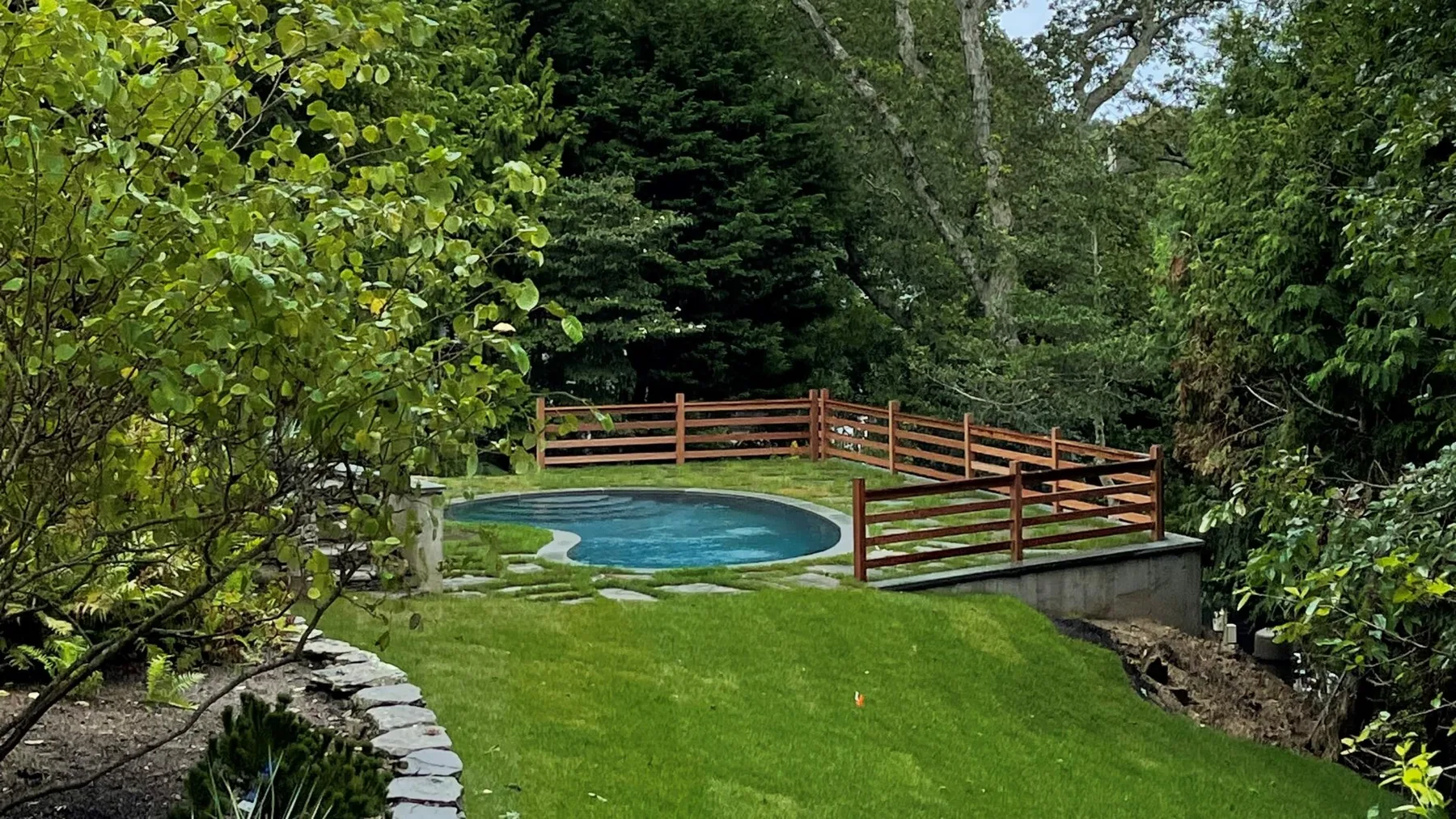A grassy garden surrounds a small pool and wooden bridge amid trees.
