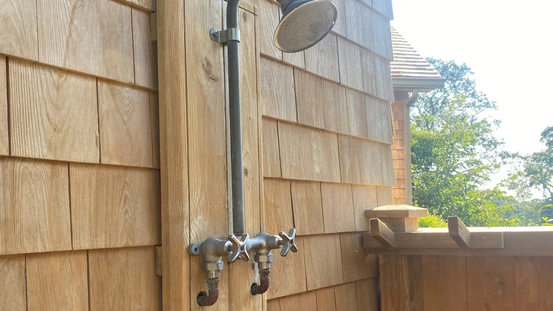 An outdoor shower is mounted against a shingled wall beside a wooden deck.