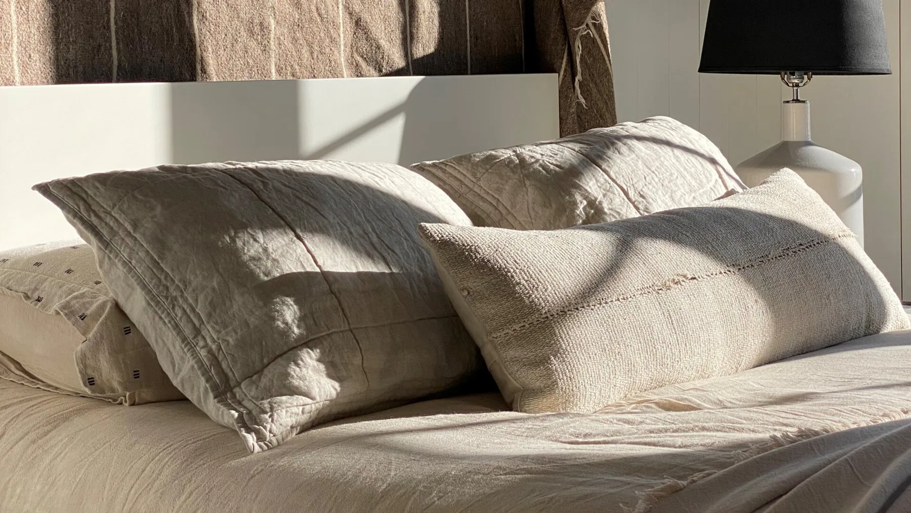 A sunlit bed with layered neutral-colored pillows and a bedside lamp.