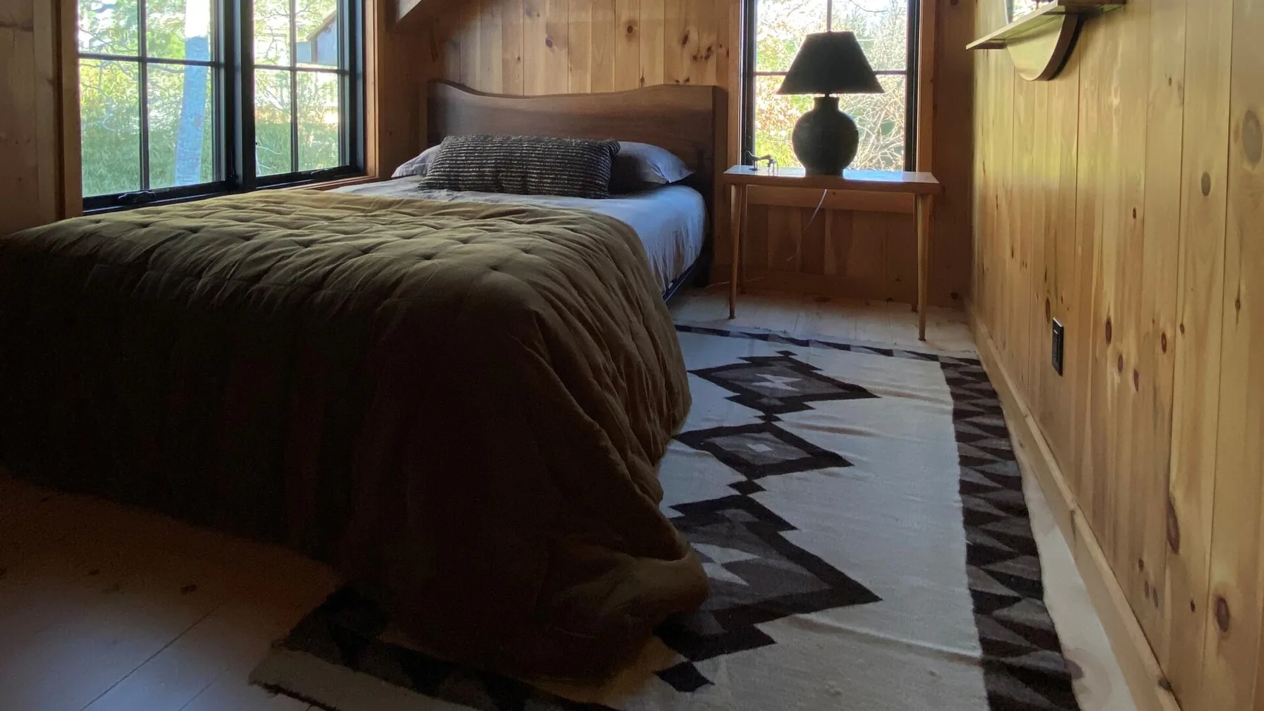 A bedroom with a bed beside large windows and a patterned rug.
