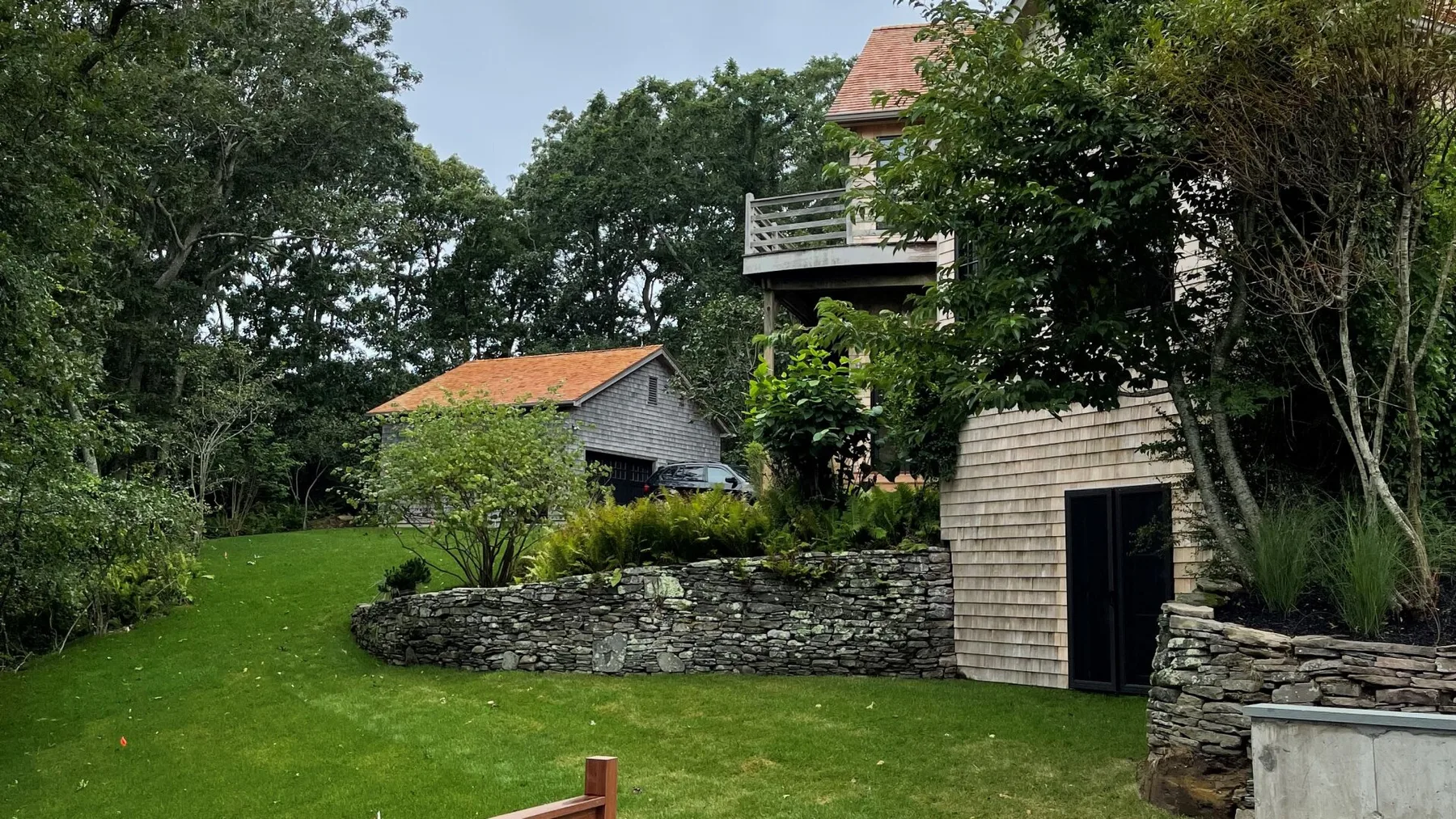 A landscaped garden with a stone retaining wall and a shingled building among trees.